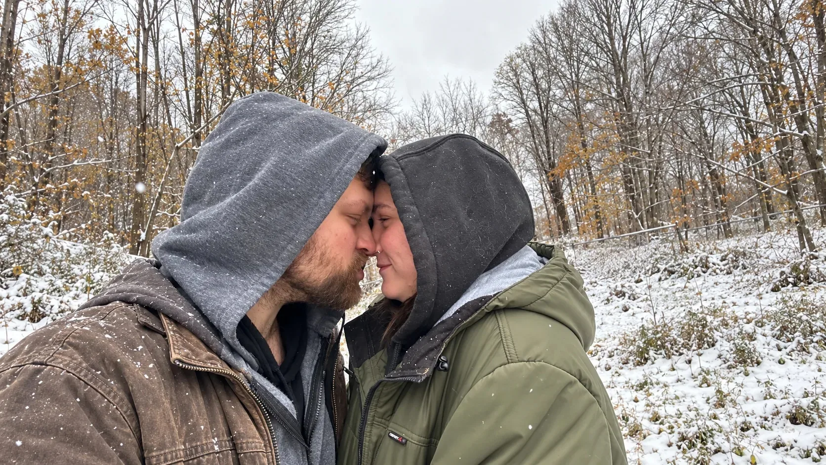 Nick and Nicole in hoods, nose to nose and smiling, as snow falls among the trees.