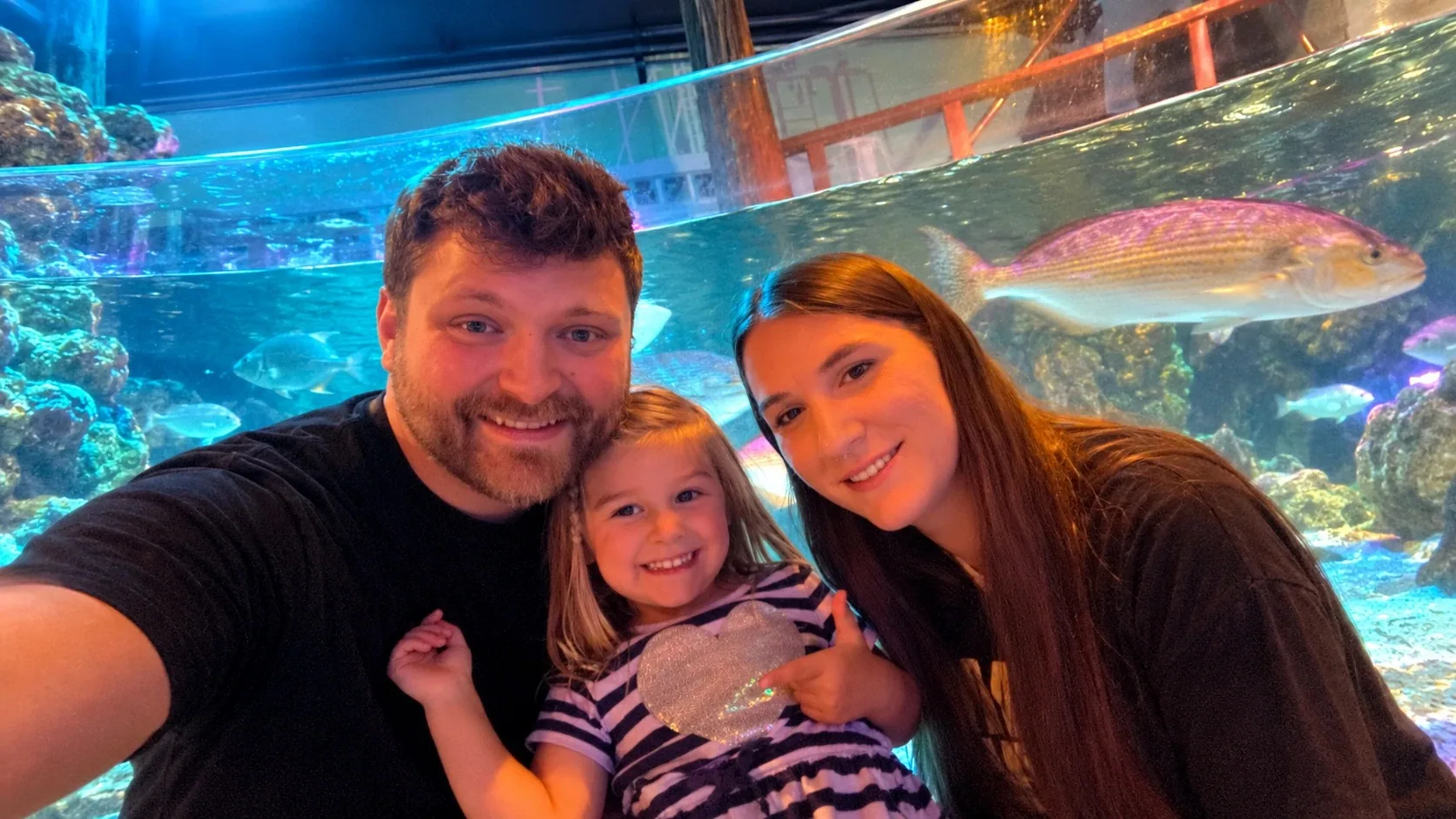 Nick, Holly and Nicole smiling together in front of a large aquarium tank with fish swimming behind them.