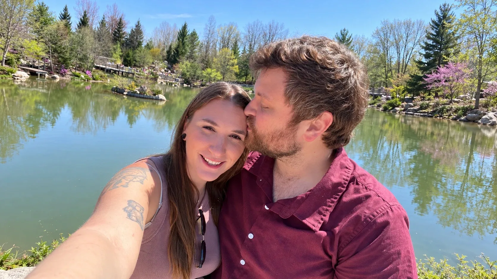 Nick kisses Nicole on the forehead as she smiles at the camera, beside a still pond with evergreens and spring blossoms on the far bank.
